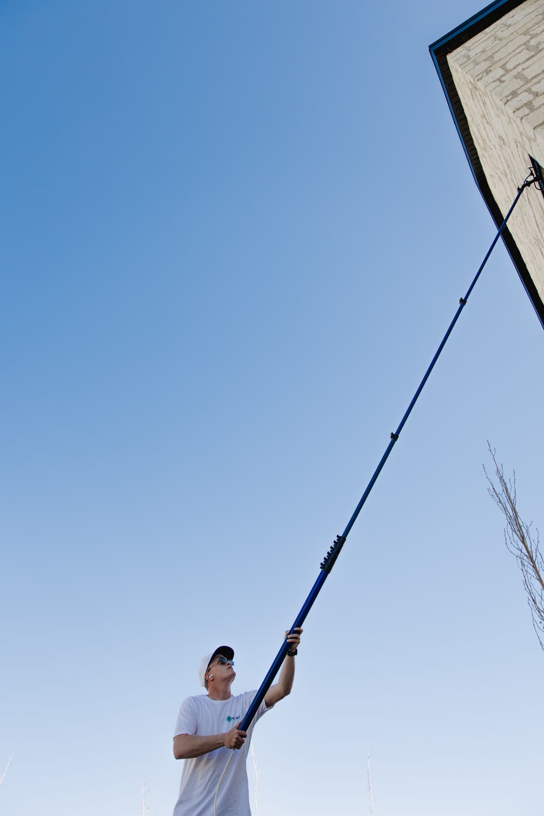 Person using a long pole to paint or clean the side of a building under a clear blue sky.