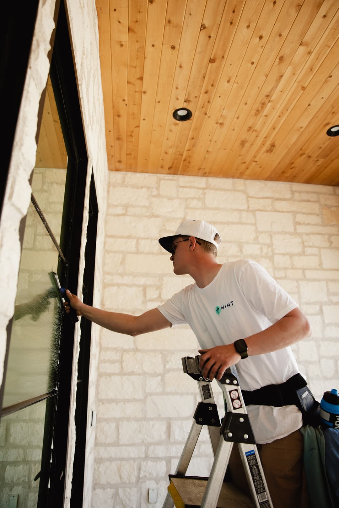 Man on ladder cleaning a window of a home, wearing a branded t-shirt and cap.
