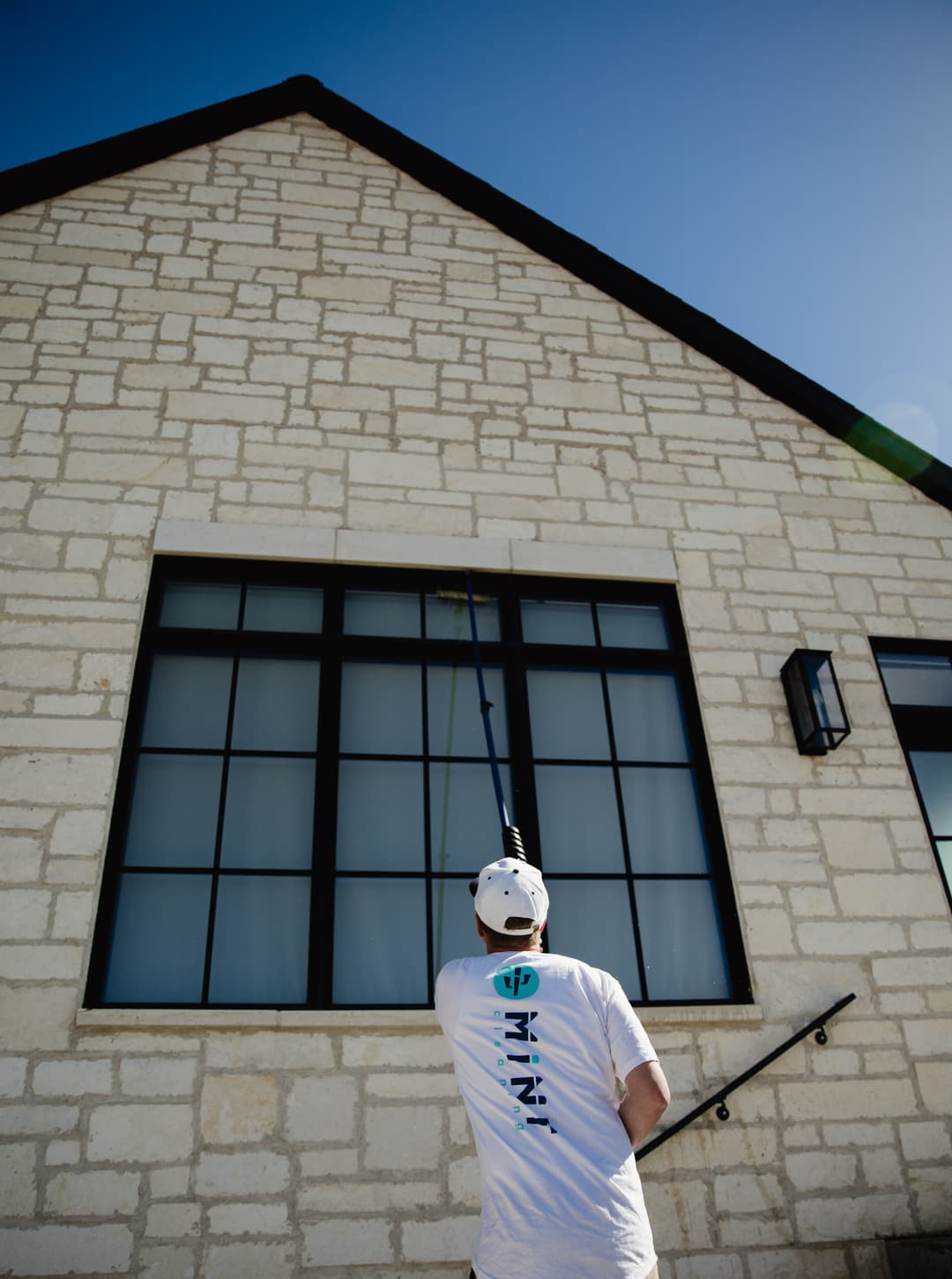 Person cleaning a large window on a modern stone building under a clear blue sky.