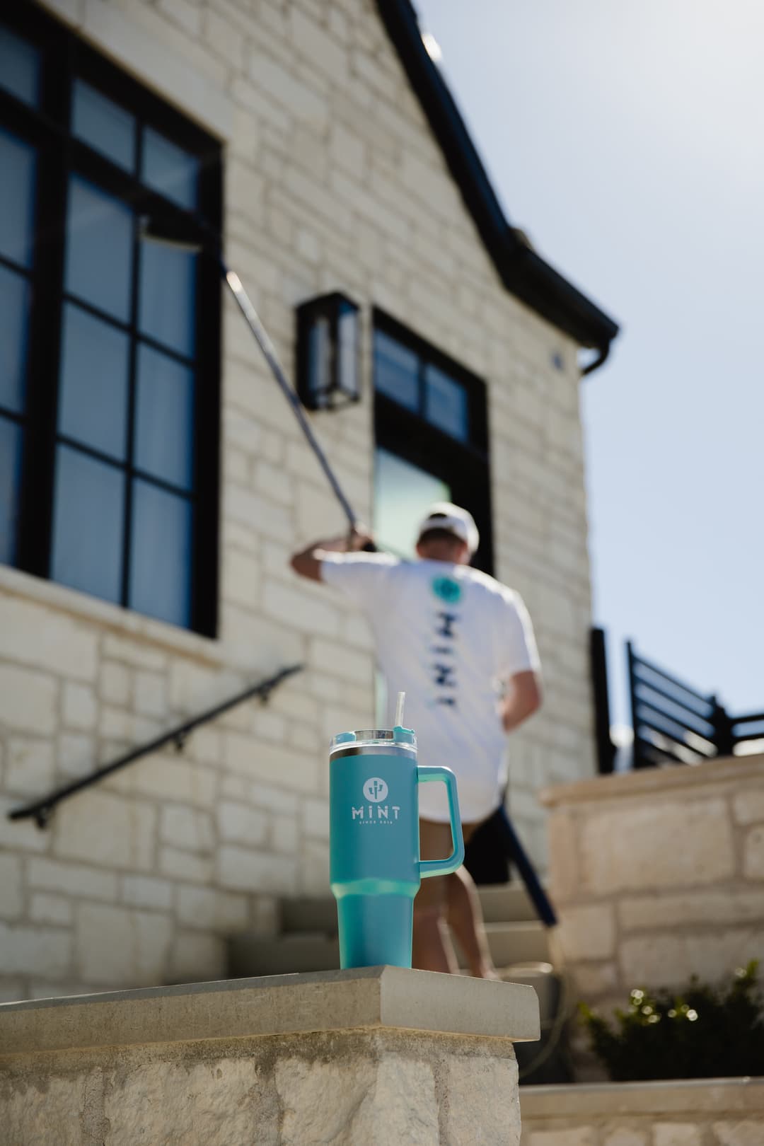 Person cleaning stairs with a Mint tumbler on the ledge, sunny day, modern stone house backdrop.