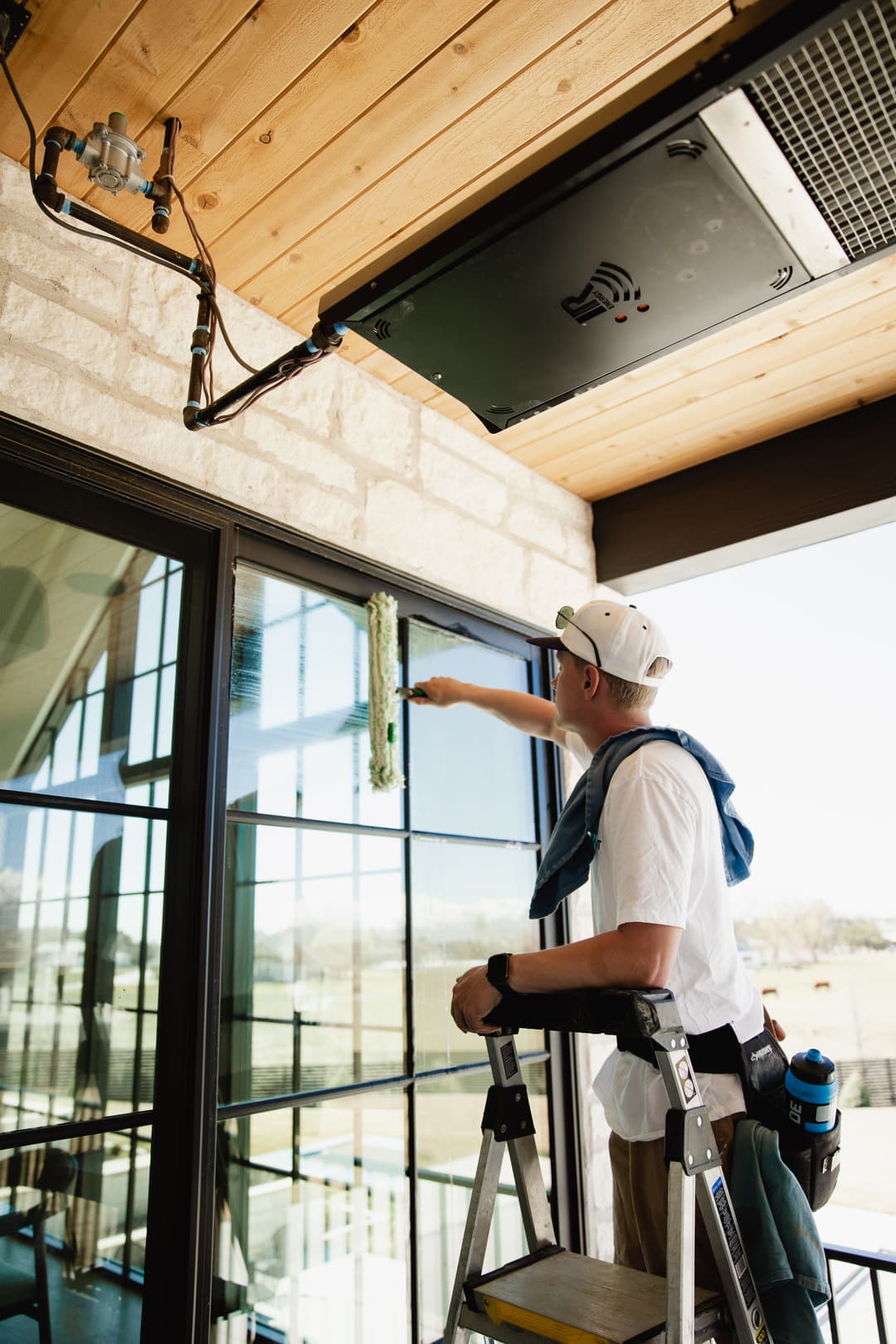 Man cleaning large windows on a ladder with tools visible, modern home exterior background.
