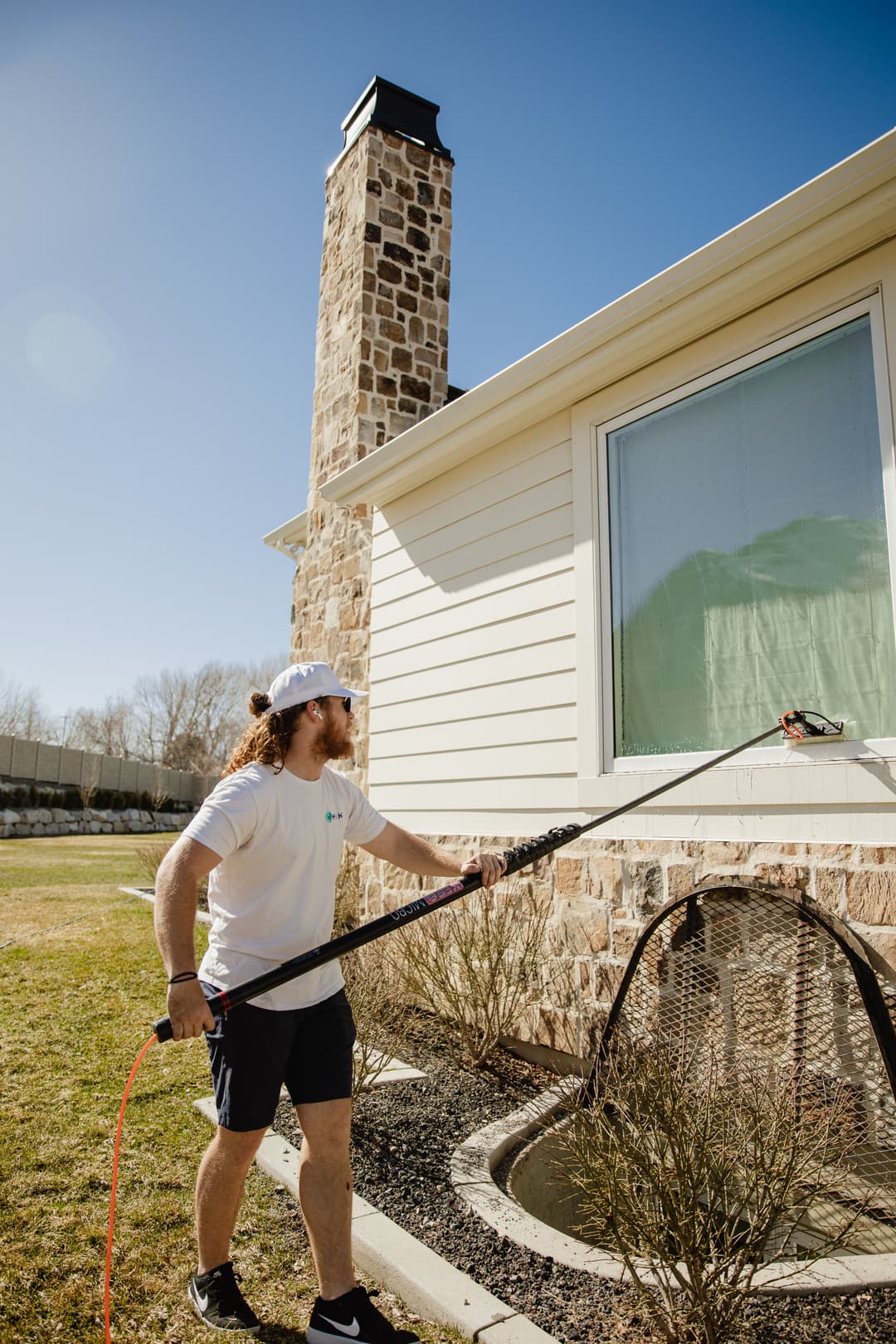 Man using a power washer on a house exterior, with a stone chimney and clear blue sky.