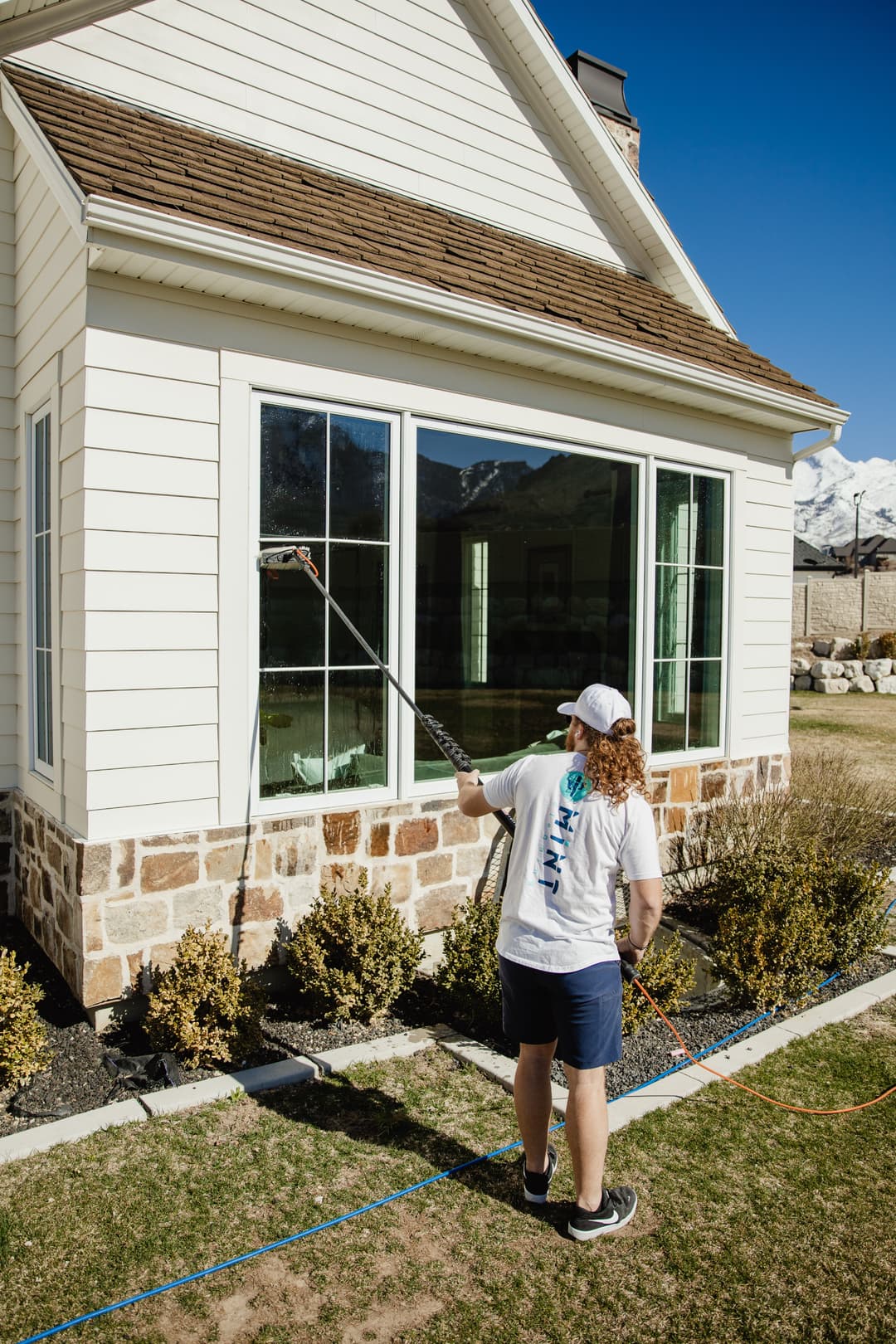 Person painting the exterior of a house with a roller, surrounded by greenery and mountains.