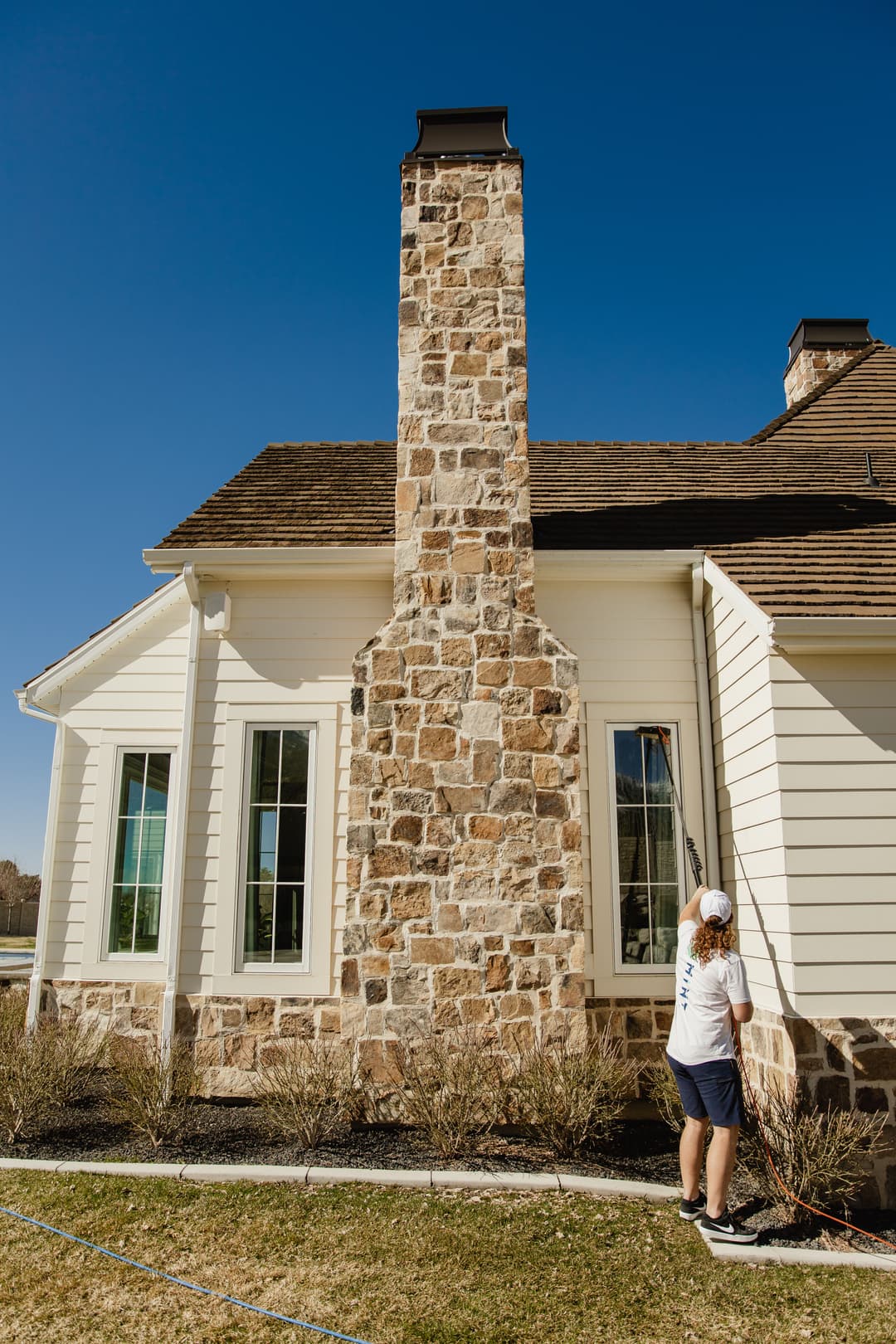Home exterior with stone chimney; person painting the wall under a clear blue sky.