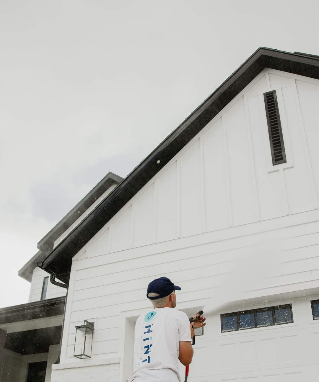 Person painting the exterior of a modern house with a paint sprayer on a cloudy day.
