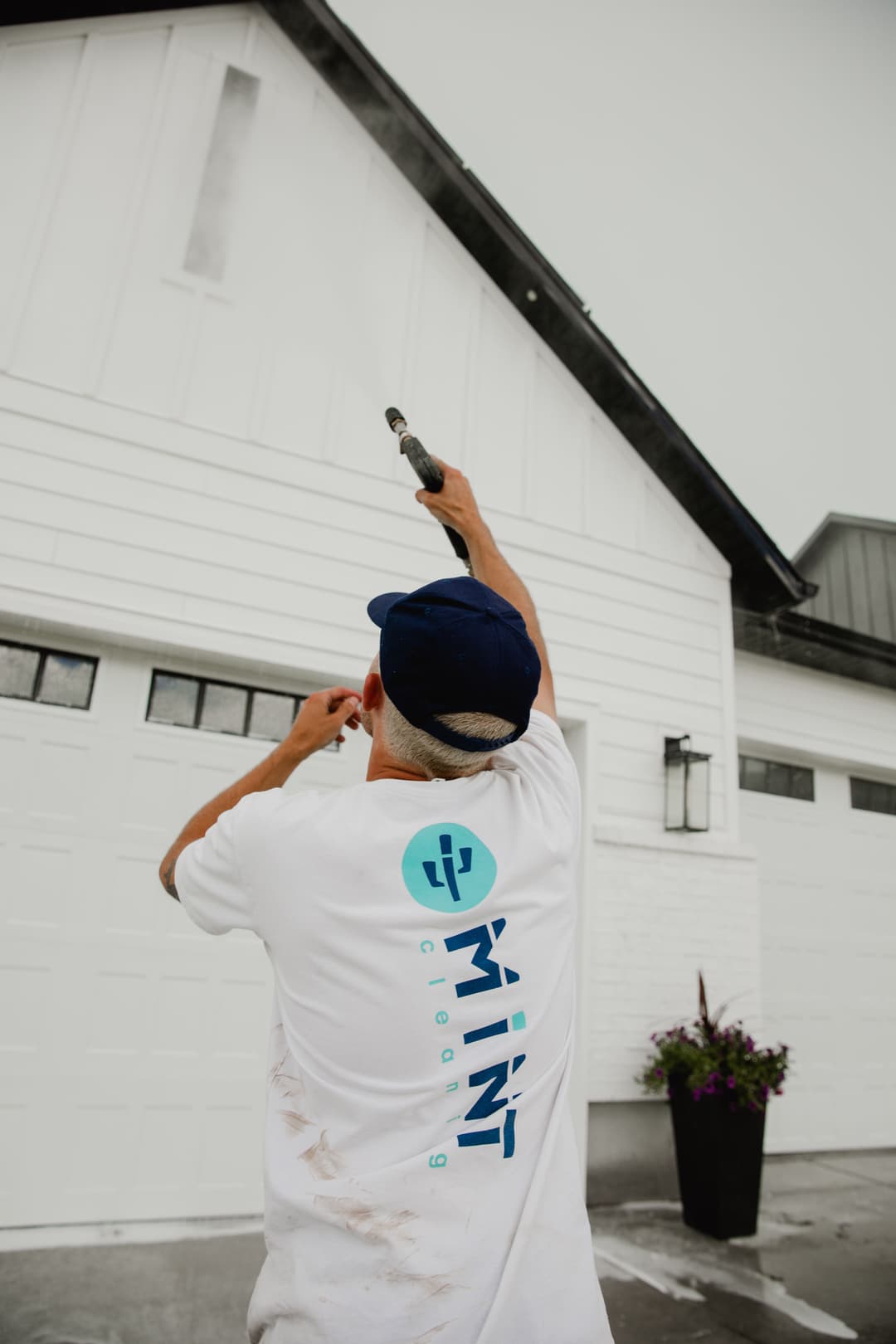 Man pressure washing a house exterior wearing a Mint Cleaning t-shirt, cloudy day.