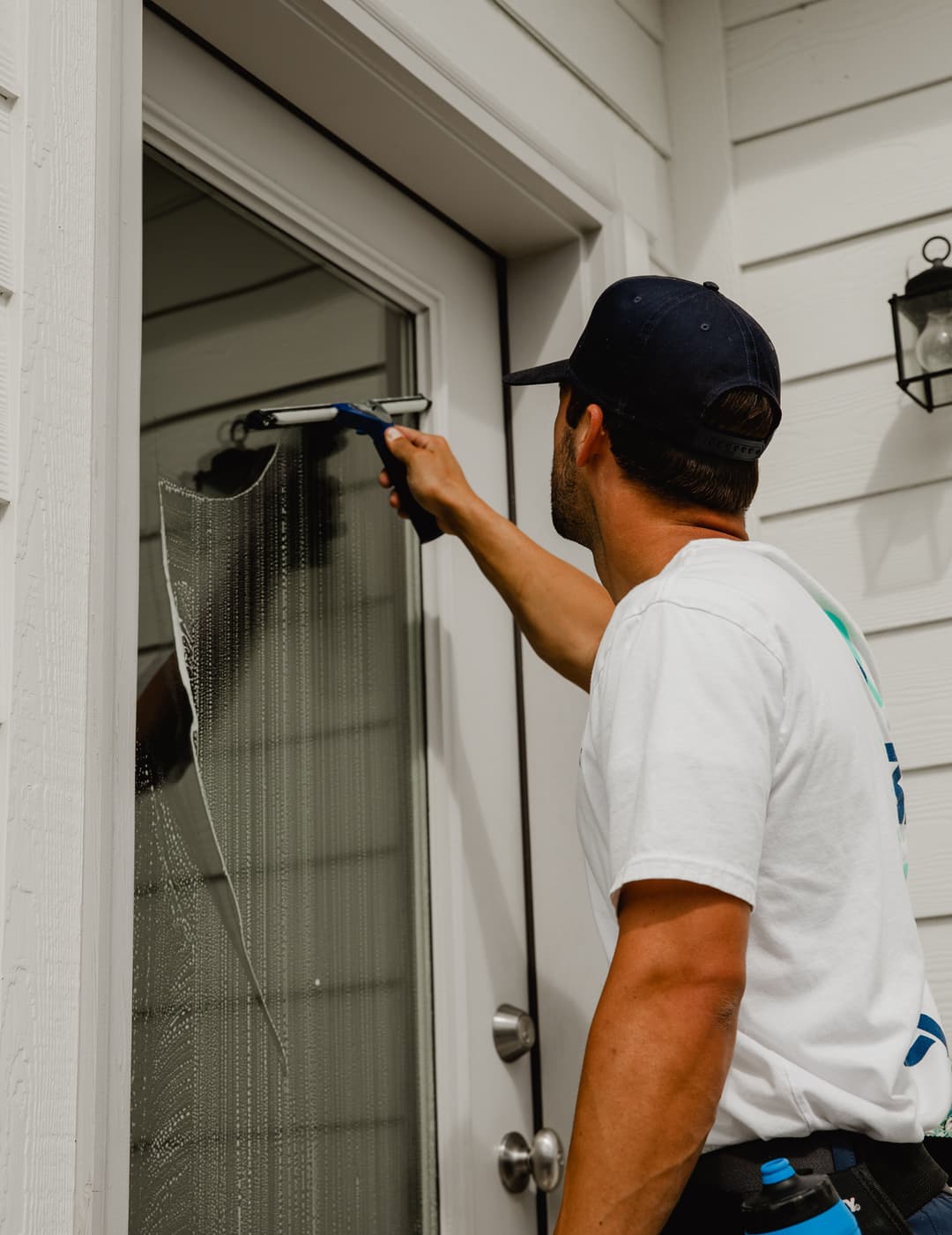 Man cleaning a glass door with a squeegee, wearing a t-shirt and cap.