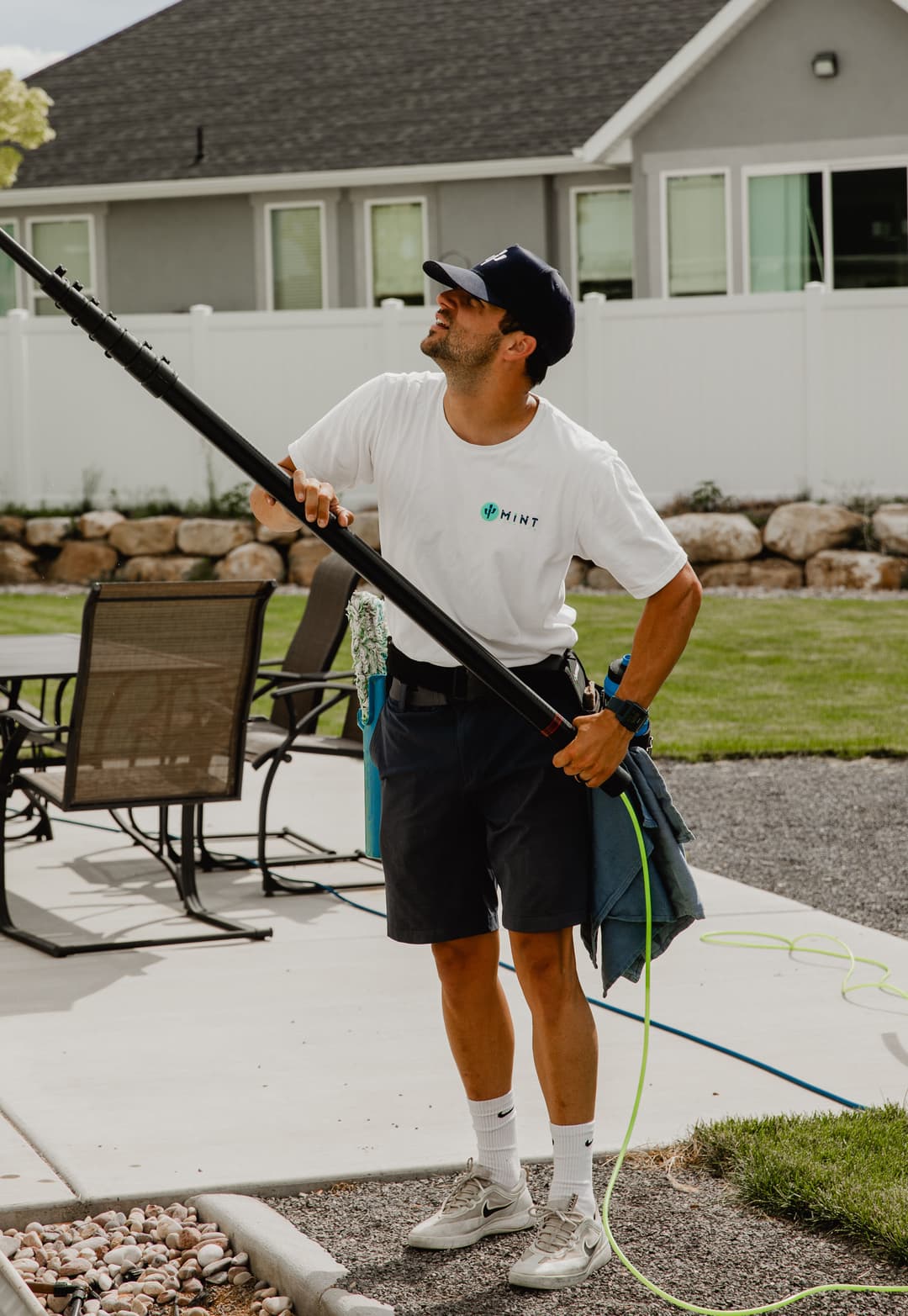 Person cleaning windows outdoors with a pole and hose in a well-maintained backyard setting.