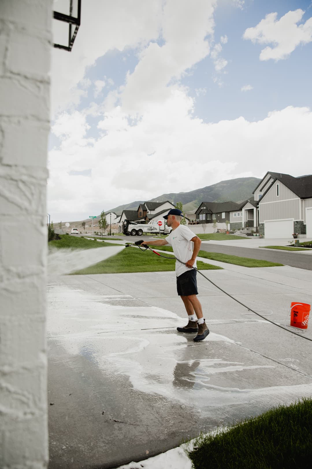 Man pressure washing driveway in residential neighborhood under cloudy sky.