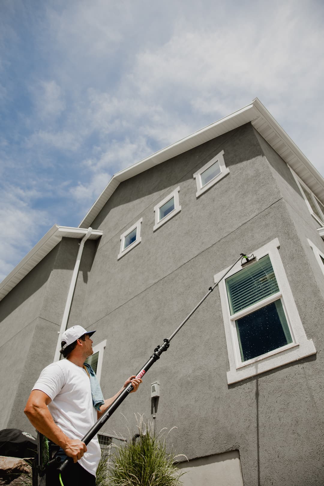 Man using a pole to clean windows on a modern gray house under a cloudy sky.