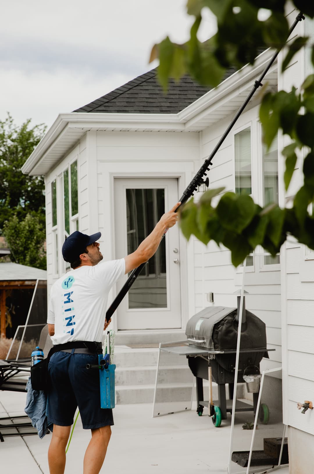 Man using a pole to clean windows on a white house with green foliage nearby.