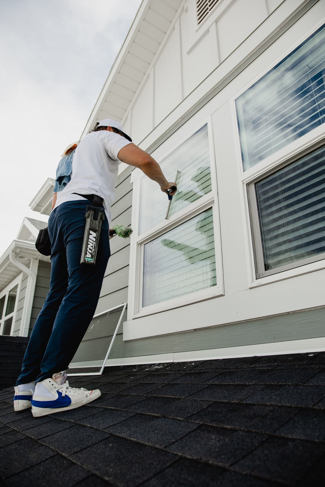 Person cleaning windows on a house roof with a squeegee and ladder, bright day.