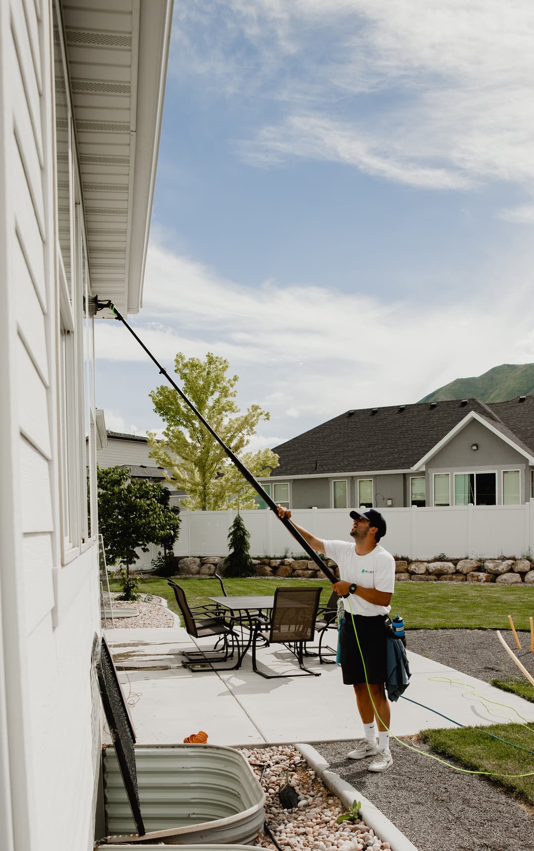 Person using a pole to clean windows on a house with outdoor furniture and mountains in the background.