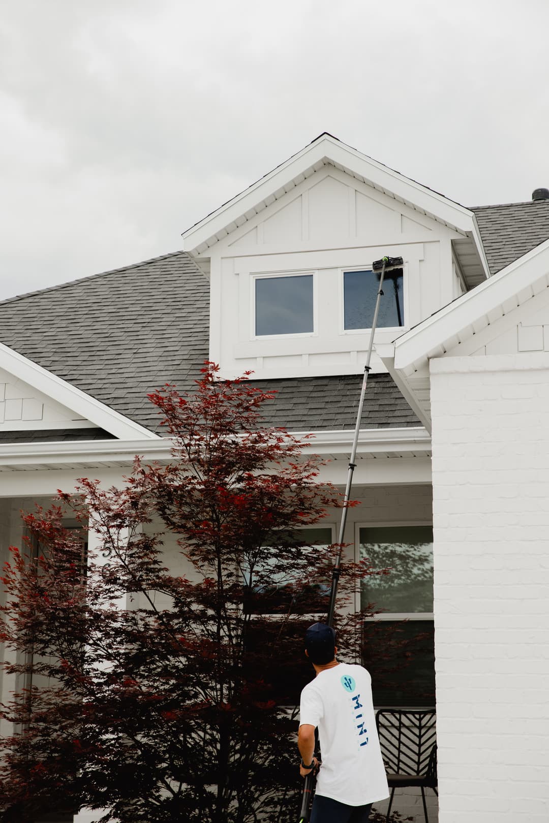 Person cleaning a house window with a long pole under a cloudy sky.