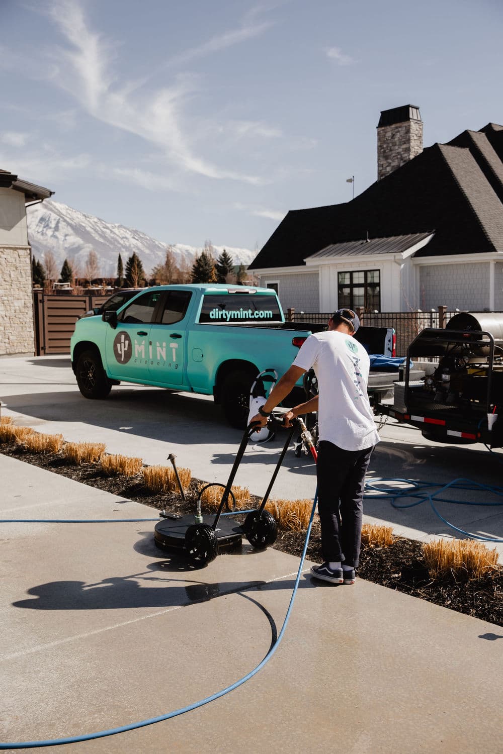 Man cleaning a driveway with a pressure washer beside a branded Mint vehicle.