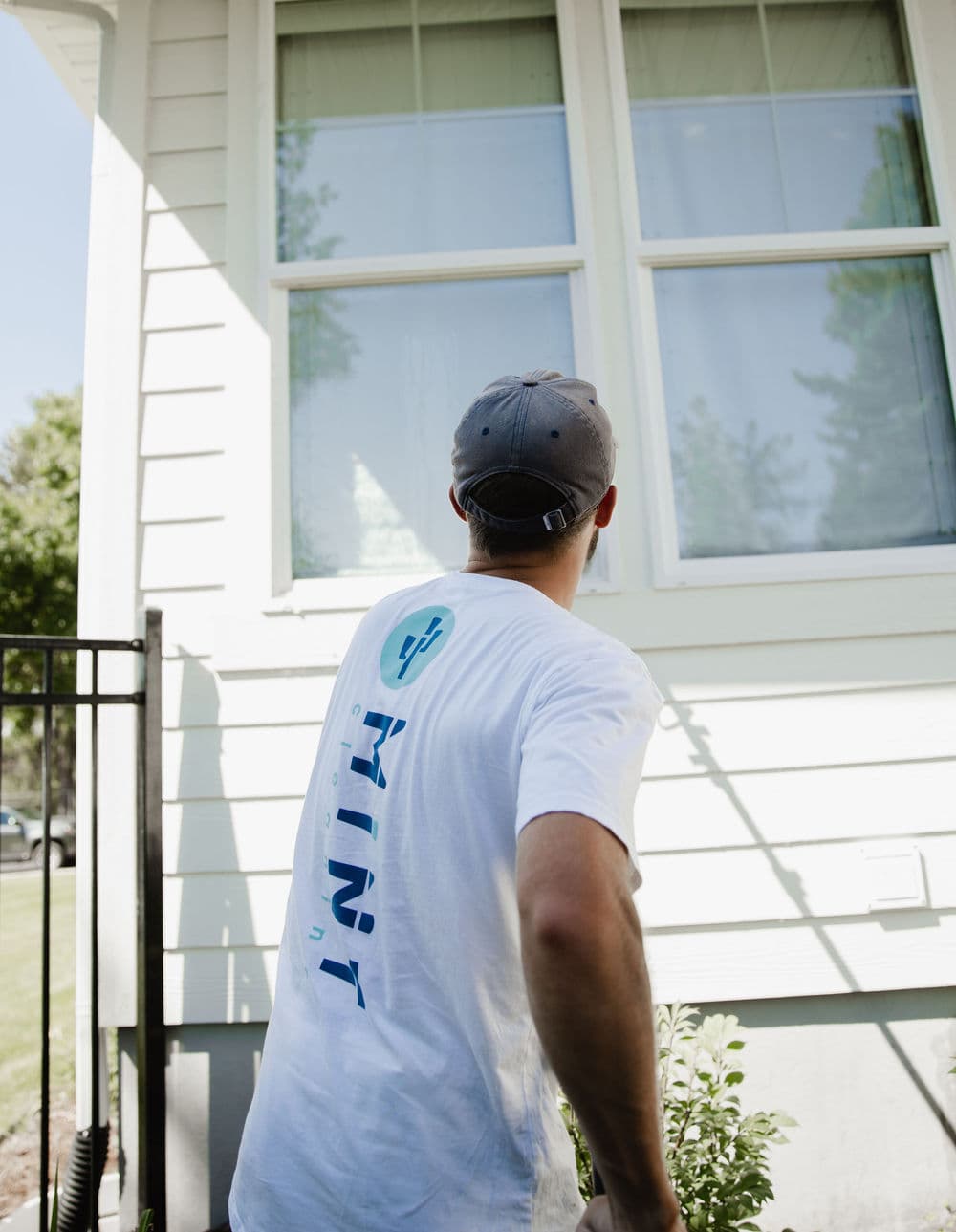 Man wearing a "Mint" shirt looking at a house window on a sunny day.