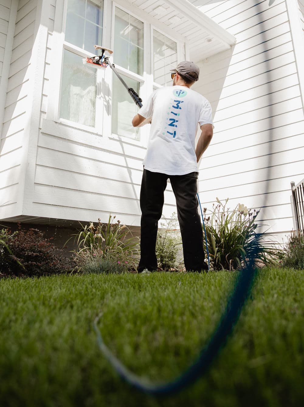 Person cleaning a window with a long pole, standing on green grass in front of a white house.