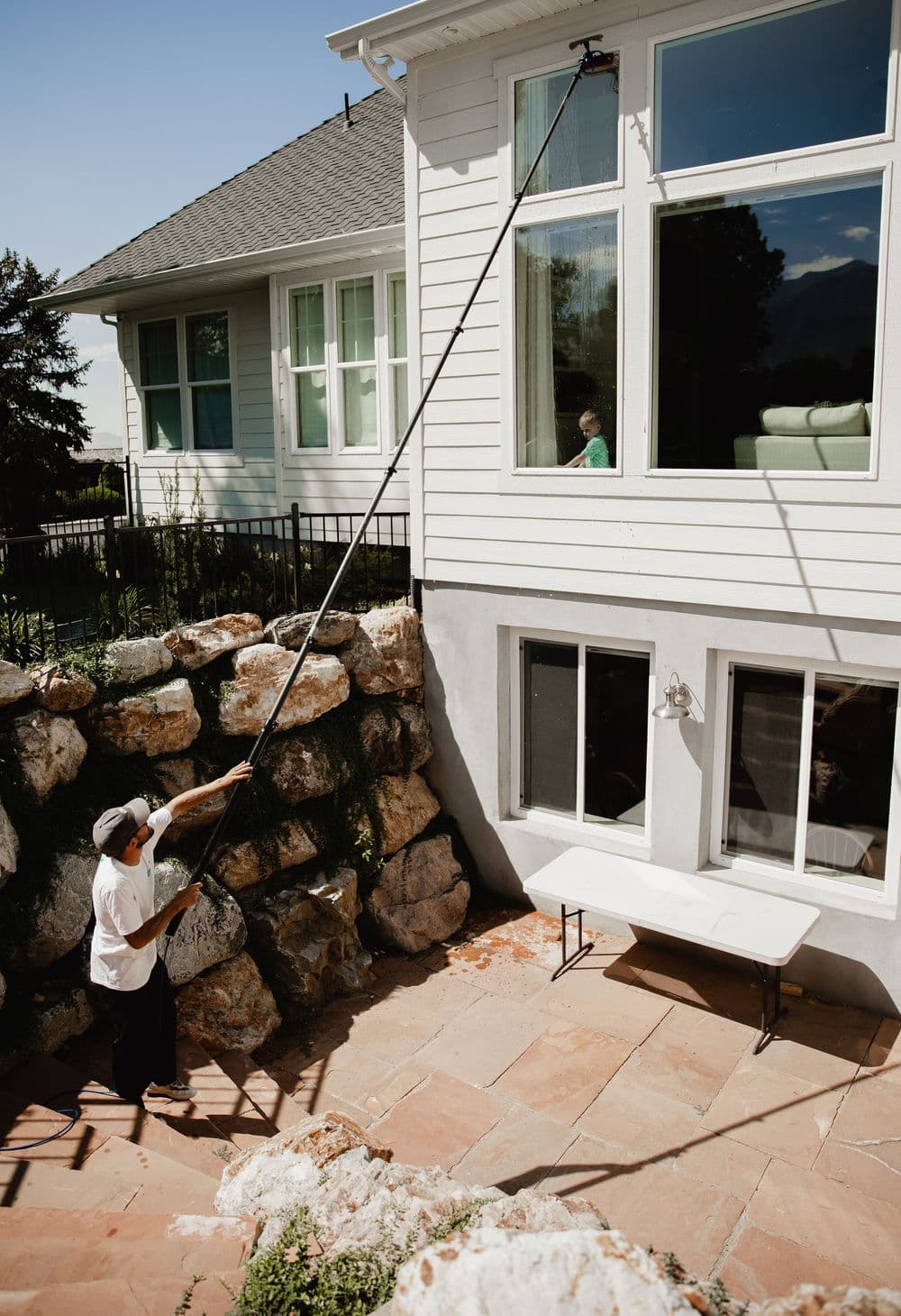 Person using a pole to clean windows on a house, with another person visible indoors.