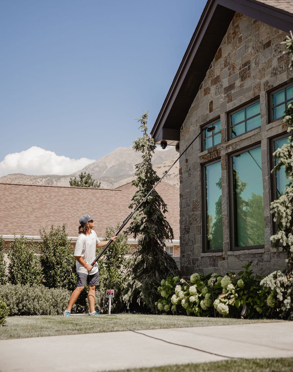 Person using a pole to clean windows on a stone house with mountains in the background.