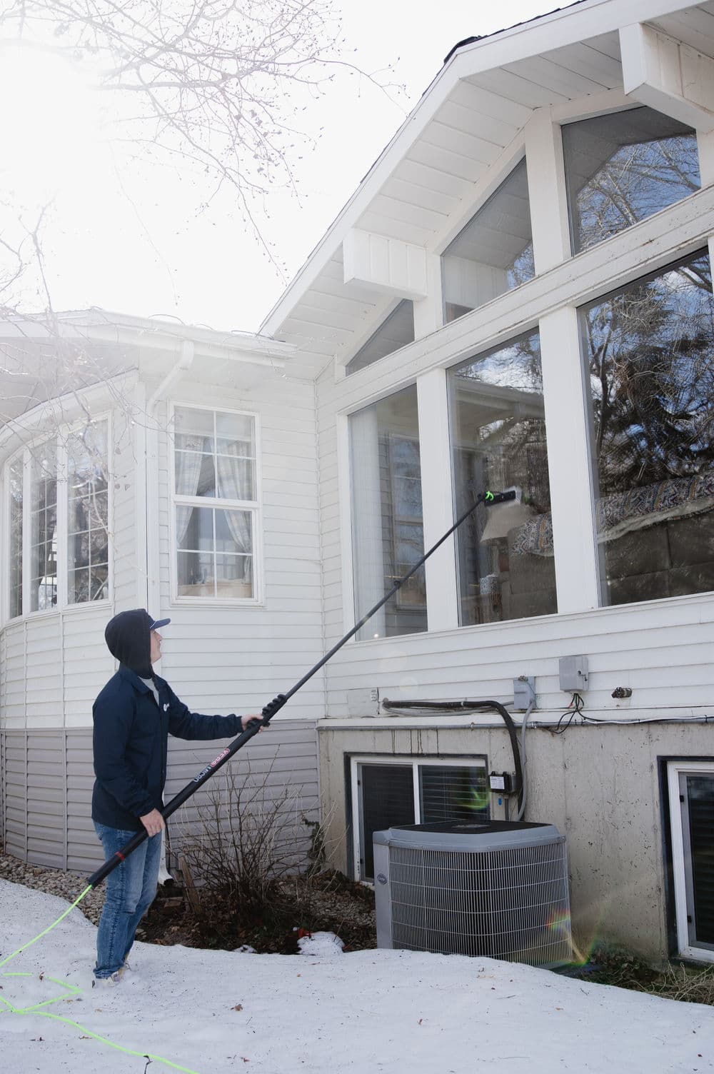 Person using a pole to clean windows of a house in winter. Snow on the ground.