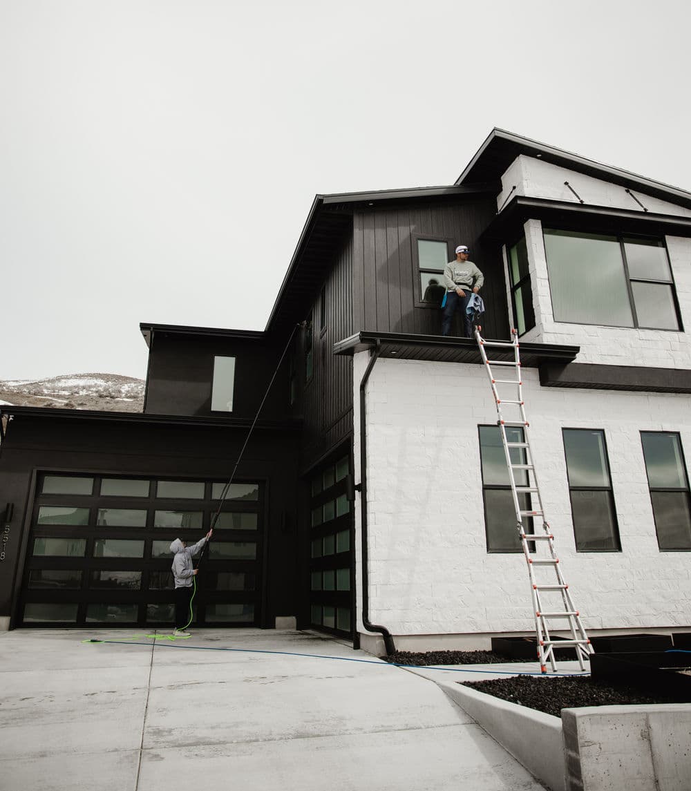 Man cleaning windows on a modern house, with a ladder and another person assisting from the ground.