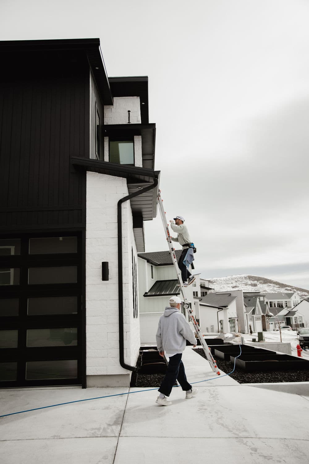 Two workers painting a modern home exterior, using a ladder on a cloudy day.