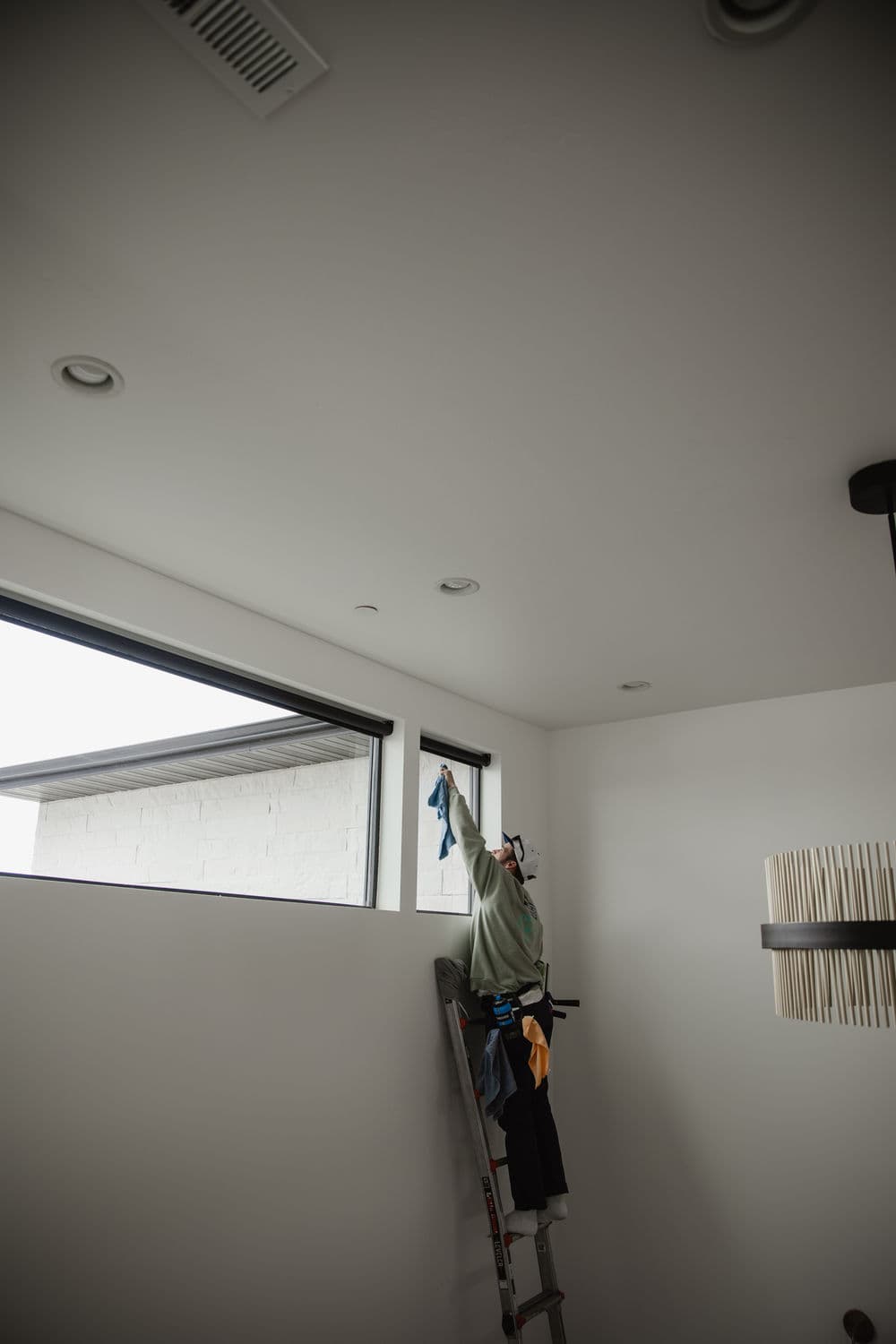 Person on ladder cleaning a window in a modern, well-lit room with white walls and fixtures.