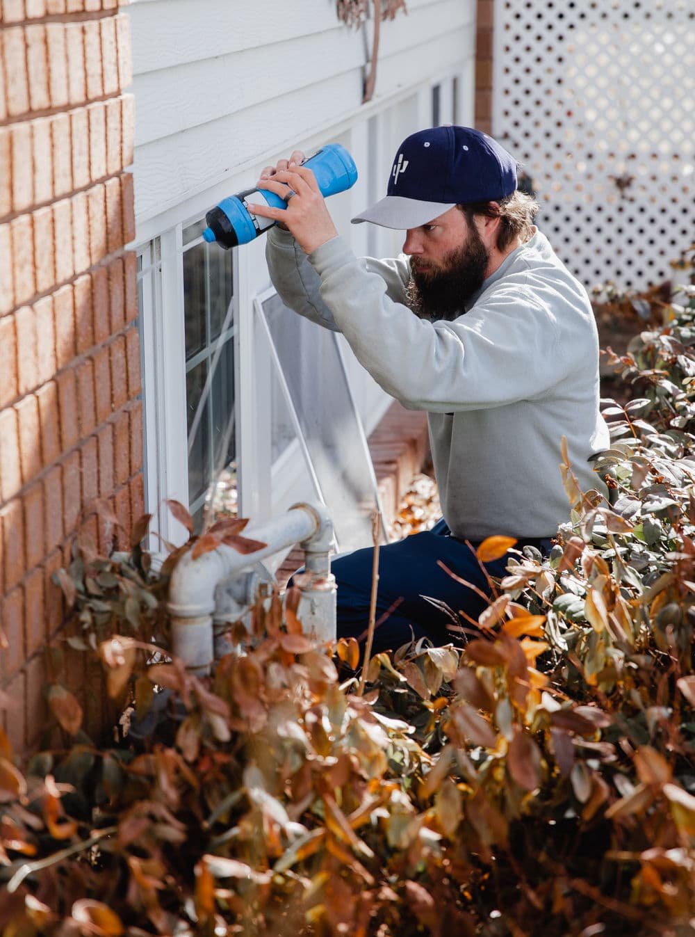 Man repairing a window with tools, surrounded by fall foliage, near brick exterior.