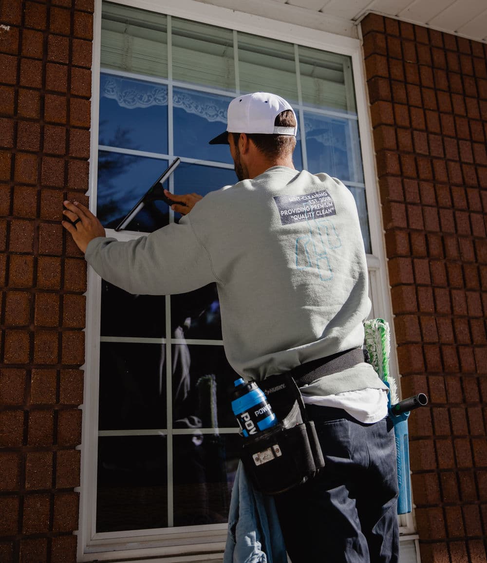 Window cleaner using a squeegee on a residential window, wearing a hat and tool belt.