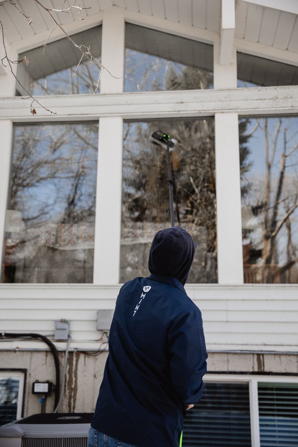 Person cleaning windows with a pole squeegee on a modern house, surrounded by trees.