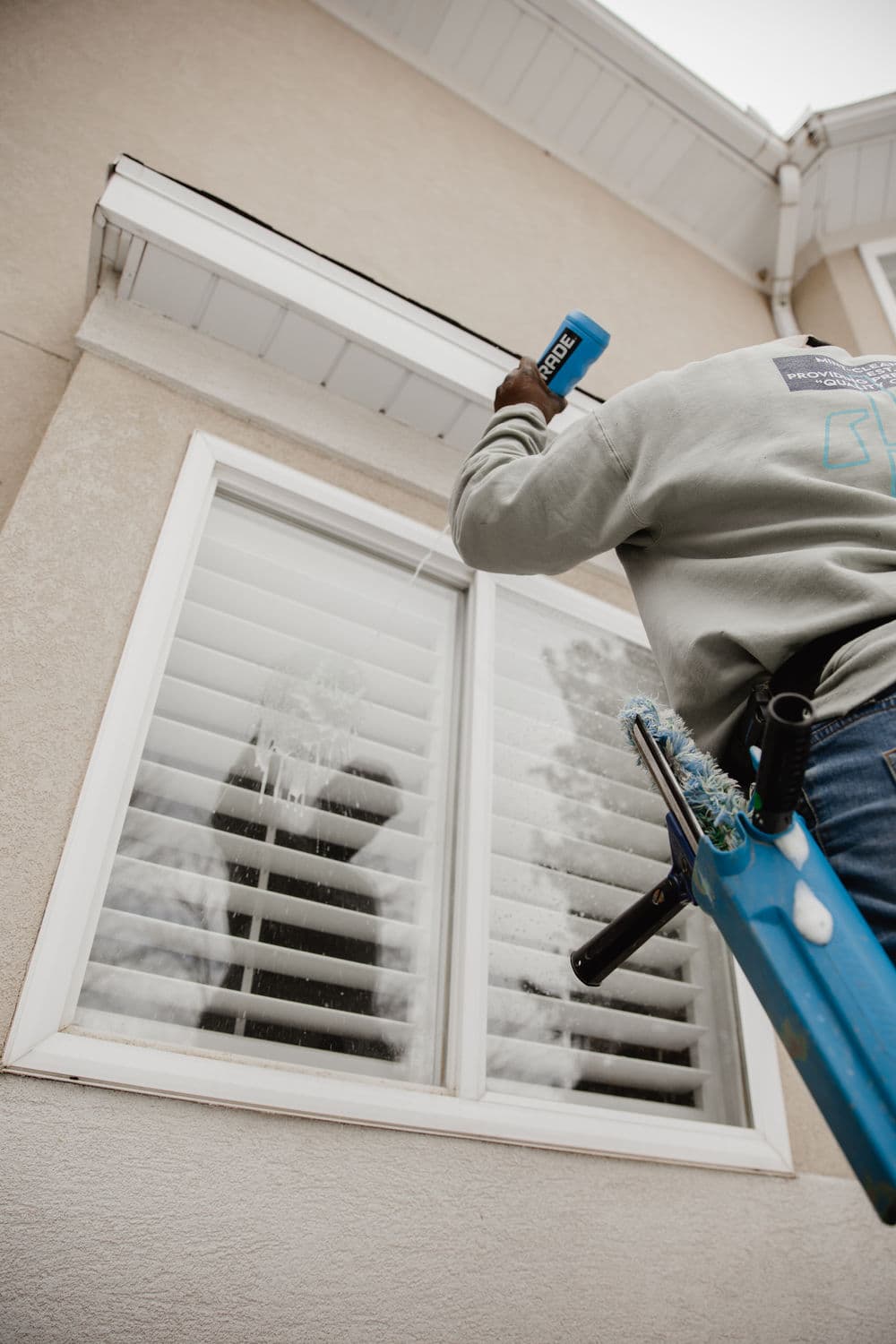 Person cleaning window blinds with spray bottle while on a blue ladder.