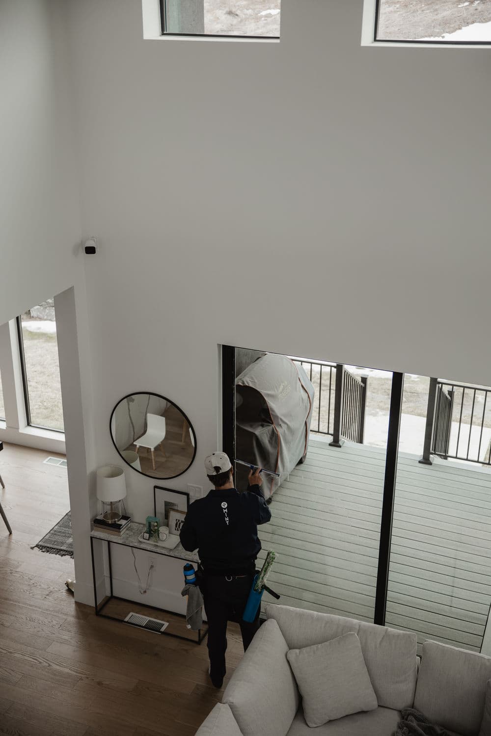 Cleaning professional dusting a modern living room with large windows and a sliding glass door.