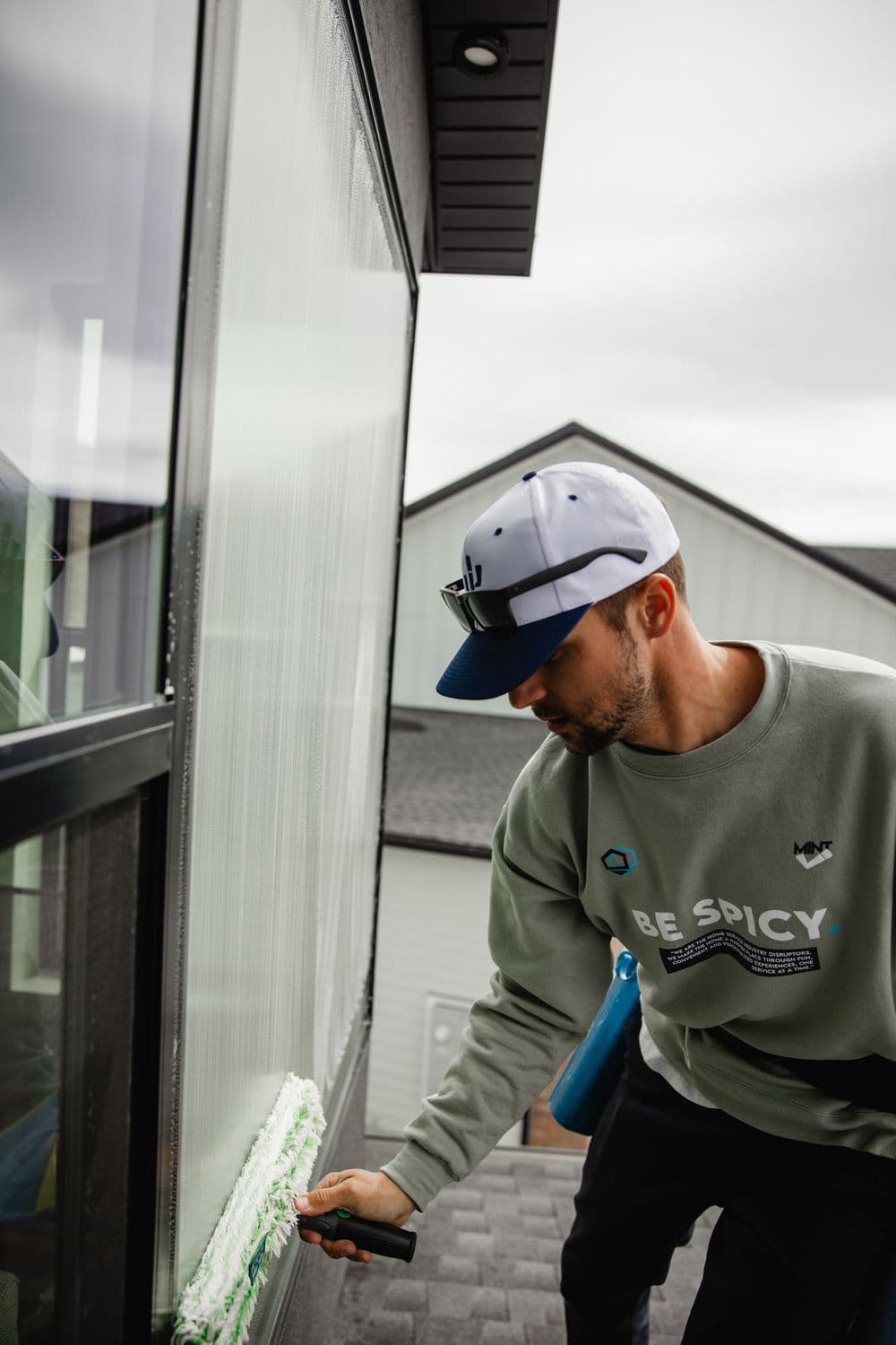 Man cleaning large window with squeegee, wearing gray sweatshirt and cap, house in background.
