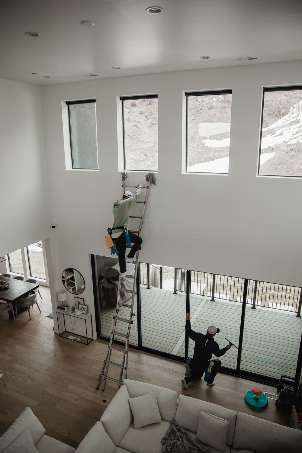 Two workers cleaning tall windows in a modern living room with a ladder and natural light.