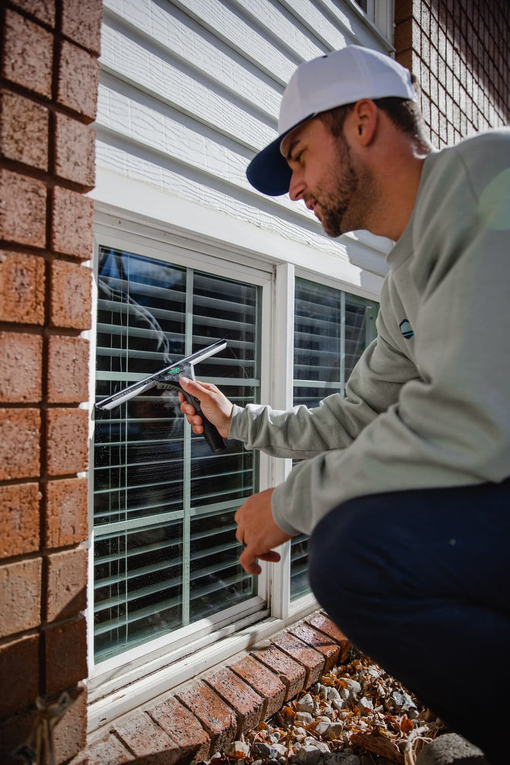 Man cleaning window with squeegee beside brick wall and white window blinds.